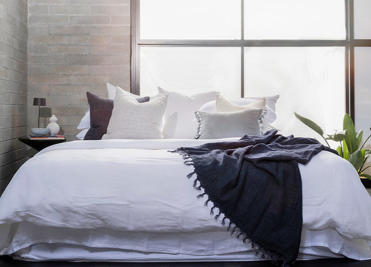 A made bed with white sheets and multiple pillows, including a bolster pillow, in a room with a window and a plant.
