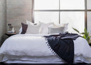 A made bed with white sheets and multiple pillows, including a bolster pillow, in a room with a window and a plant.
